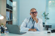 © amnaj - Senior male doctor sitting at desk with hand on chin, exuding confidence and professionalism in his medical office
