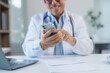 © amnaj - Smiling doctor using smartphone in medical office with laptop and documents on desk