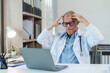 © amnaj - Senior doctor having a bad headache while working on a laptop in his bright office