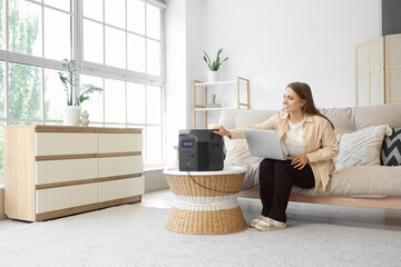 Young woman charging laptop with portable power station on sofa at home