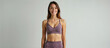 © Roman - A young woman with long brown hair is smiling at the camera. She is wearing a purple sports bra and purple leggings. She is standing against a white wall. She has a toned physique and is looking confi