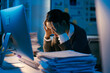 © Parichat - Young businesswoman is having a headache and feeling stressed while working late at night in the office, surrounded by stacks of paperwork and using a computer