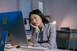 © Parichat - Exhausted asian businesswoman sitting in a dimly lit office at night, looking bored and tired while using a computer, captures the struggle of work-life balance and corporate stress