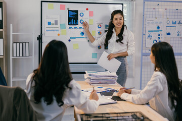 Fototapete - Asian businesswoman leads a meeting in the office, presenting charts and statistics on a whiteboard to her colleagues who are taking notes