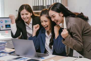 Fototapete - Three young asian businesswomen are celebrating a successful project while looking at a laptop screen in a modern office, raising their fists in excitement and expressing joy