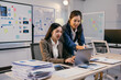 © PaeGAG - Two young businesswomen collaborating on a project in a modern office, analyzing charts on a laptop surrounded by paperwork and a whiteboard with graphs