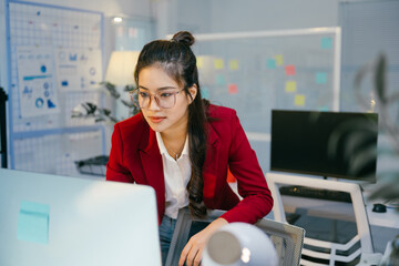 Fototapete - Young asian businesswoman wearing eyeglasses and red jacket concentrating on working with desktop computer in modern office, with charts and graphs on the wall in the background