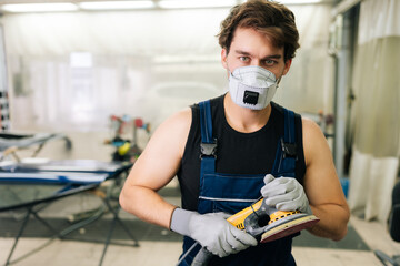 Mechanic male using machinery car polishers maintenance to remove marks repair. Portrait of muscular expert car paint technician in face mask holding car polisher to polish of car with refinement.