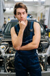© dikushin - Vertical portrait of thoughtful mechanic male in uniform looking at camera not knowing what disrepair during maintenance car in repair workshop. Doubtful repairman trying to solve problem in garage