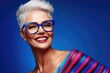 © Konstiantyn Zapylaie - Confident senior woman with stylish gray hair and bold glasses smiling against blue background