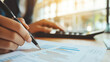 © Jirapron - Person analyzing financial data with pen and calculator in soft natural light, focusing on graphs and charts for investment decisions