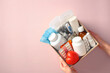 © photoguns - Female hands holding a first aid kit box with essential medical supplies on a pink background. Medical donations, first aid kit, home delivery of medications, online pharmacy concept.