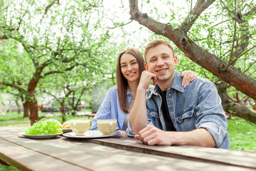 Wall Mural - young couple in love in denim clothes sit in the garden at a wooden table on a picnic in the summer and look at the camera, a guy and a girl on a date drink coffee with grapes in the park