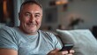 © peppastocker - An older man with short hair and wearing a casual T-shirt sits on a couch, using a smartphone and smiling at the camera, creating a comfortable and relaxed atmosphere.