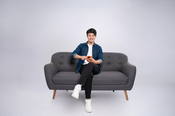 full body image of young asian man using phone sitting on sofa on white background