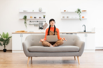 Wall Mural - Caucasian woman sitting in lotus pose meditating during remote work at home with laptop on sofa. Bright and modern living space with calming atmosphere.