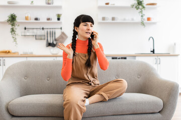 Wall Mural - Young Caucasian woman sitting on sofa in modern kitchen having phone call. Wearing casual orange and brown outfit, she gestures expressively, engaged in conversation.