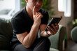 © bongkarn - A close-up of a thoughtful Asian man using his smartphone on a sofa indoors, reading a chat.