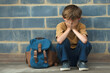 © ChaoticDesignStudio - Young boy sitting on the floor in distress beside a blue backpack against a brick wall