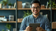 © AGCreative - smiling Asian man holding tablet in modern office setting, surrounded by plants and bookshelves. He appears confident and engaged