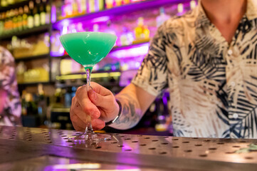 Wall Mural - A close-up of a hand holding a blue cocktail in a martini glass, with a blurred bar background. The vibrant setting highlights themes of socializing, nightlife, and beverages