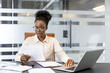 © Liubomir - Young african american business woman focused on reading documents at desk in modern office. Professional environment with laptop and paperwork showcasing concentration and productivity.