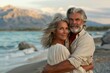 © neatlynatly - A senior couple stands on a beach, embracing and smiling warmly at the camera with mountains and ocean in the background, creating a romantic and peaceful scene.