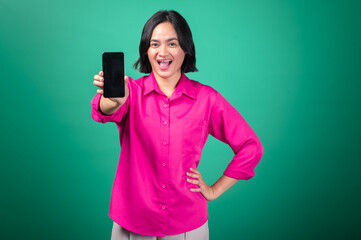 An Asian woman in a vibrant pink shirt stands confidently with one hand on her hip, holding up a smartphone with a smile against a green background.