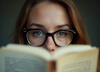 © fotoluigi868 - Young woman in eyeglasses hides behind a book while gazing directly at the camera with a subtle introspective expression