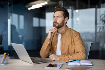 Wall Mural - Mature businessman in modern office environment sitting at desk with laptop, phone, and open notebook, conveying thoughtful expression. Professional setting highlights business and work.