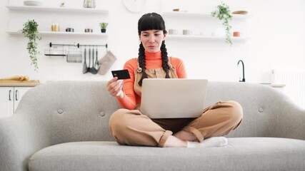 Wall Mural - Caucasian woman in twenties uses laptop on sofa for online shopping. She holds bank card, dressed casually in bright interior. Image conveys convenience, technology, and modern lifestyle.