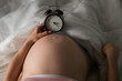 © Михаил Решетников - A pregnant woman lies in bed and holds an alarm clock. Close up of the belly.