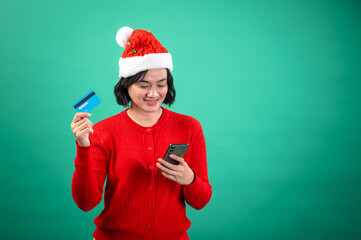 Asian woman in a red sweater and Santa hat is excitedly holding a blue credit card in one hand and a smartphone in the other against a green background, symbolizing festive holiday shopping