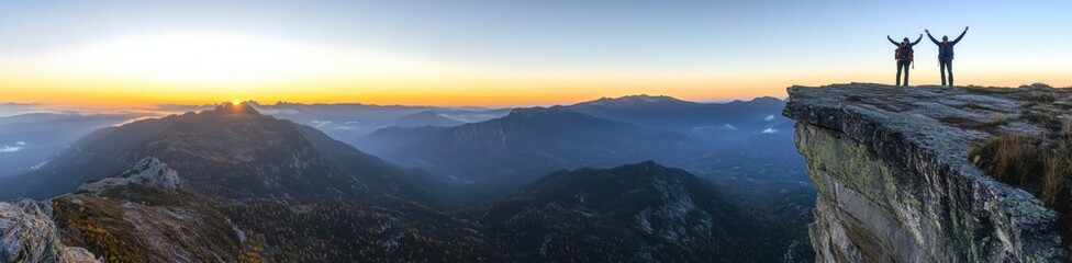 Wall Mural - A panoramic view of two hikers standing on the edge of an alpine cliff, arms raised in joy as they reach their goal at sunrise, with misty mountains and a clear sky in the background Generative AI