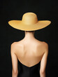 © Vilma - A woman with her back to the camera, wearing a large straw hat and a black swimsuit. The background is dark, emphasizing her silhouette and the hat's wide brim.
