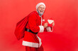 © khosrork - Side view of smiling elderly man with gray beard in santa claus costume standing with bag of presents and holding letter with Christmas congratulations.
