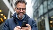 © Pinklife - Middle-aged man with glasses happily checks his smartphone while standing in an urban environment, showcasing modern technology's role in everyday life.