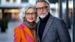© Pinklife - An older couple wearing colorful winter coats and scarves stand close together, posing for a portrait in an urban setting, exuding warmth and elegance.