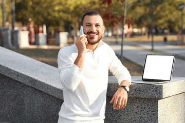 Wall Mural - young man with laptop talking by mobile phone in park