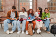 © Jose Calsina - Diverse Group of College Students Engage with Their Smartphones While Studying Together on Campus Bench, Mixing Social Media and Academic Work Against Historic Brick Architecture