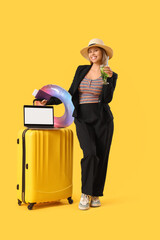 Wall Mural - Young businesswoman with travel accessories and laptop waiting for summer vacation on yellow background