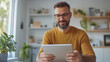 © NaphakStudio - man with beard is sitting at table, smiling while using tablet in bright, modern room filled with plants and decor. His relaxed demeanor suggests enjoyment and engagement with device.