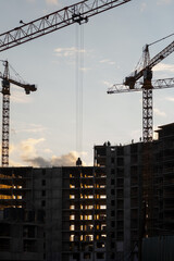 Fototapete - Cranes operate at a construction site during sunset, with workers visible on the building's framework as daylight fades