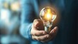 © Phongsri - A close-up of a human hand gently holding a glowing lightbulb against a soft-focus background, symbolizing ideas, innovation, and inspiration in a creative context.