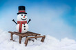 © fogaas - A happy snowman in a top hat and scarf, arms up and ready for winter fun! Posing with a sled on a snowy background and bright blue sky.