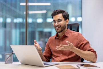 Smiling Hispanic businessman sitting at desk using laptop for video call in modern office setting. Glasses and professional attire emphasize business context. Concept of communication, technology