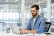 © Liubomir - Confident man with glasses using laptop in bright office setting. Engaged in work, showcasing productivity and focus in modern workspace with natural light and clear glass walls.