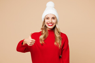 Poster - Merry fun young woman wear red warm cozy knitted sweater hat posing showing thumb up like gesture isolated on plain pastel light beige background. Happy New Year celebration Christmas holiday concept.