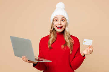 Poster - Merry young woman in red sweater hat posing use laptop pc computer hold credit bank card shopping online isolated on plain light beige background Happy New Year celebration Christmas holiday concept