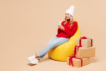 Canvas Print - Full body smiling merry young woman she wears red warm cozy sweater hat posing sit in bag chair isolated on plain pastel light beige background. Happy New Year celebration Christmas holiday concept.